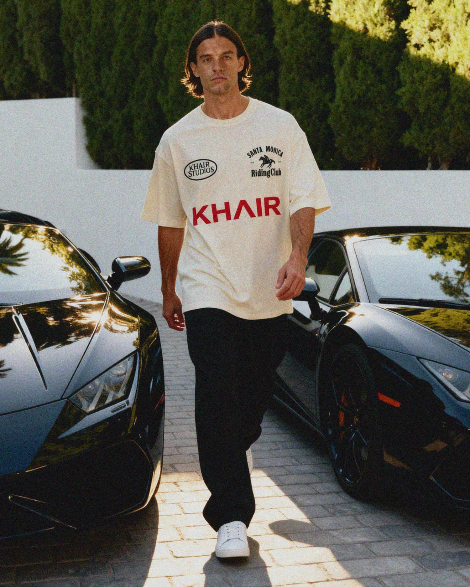 Man wearing a white KHAIR Santa Monica Riding Club t-shirt with logos and text, standing between two black luxury cars.
