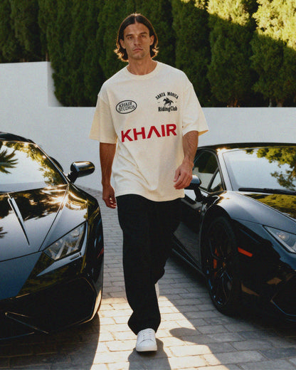 Man wearing a white KHAIR Santa Monica Riding Club t-shirt with logos and text, standing between two black luxury cars.