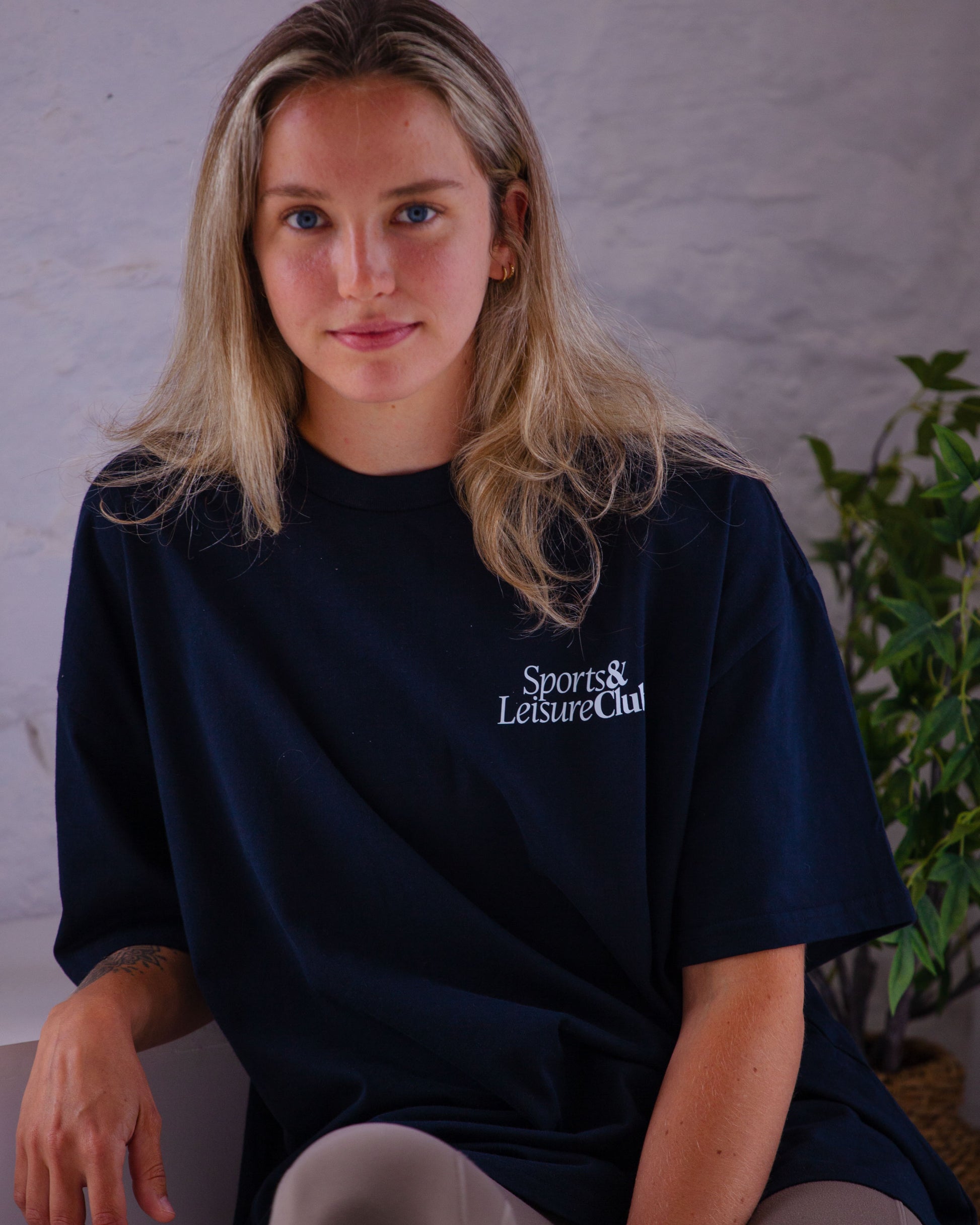 Woman wearing navy oversized Sports & Leisure Club t-shirt with white print, seated indoors in relaxed setting