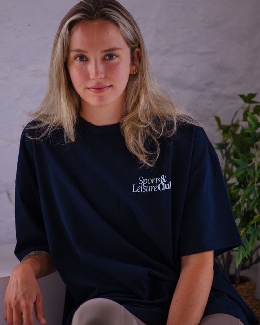 Woman wearing navy oversized Sports & Leisure Club t-shirt with white print, seated indoors in relaxed setting